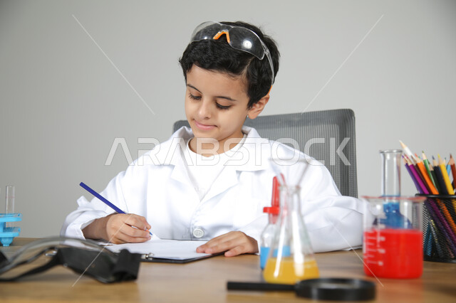 Embodying children's future professions since childhood, passion to reach the dream of studying pharmacy in the future, chemical experiments for young scientists, close-up portrait of a Saudi Arabian Gulf boy wearing a medical coat sitting behind a desk with analysis bottles in front of him and recording notes with concentration, gray background
