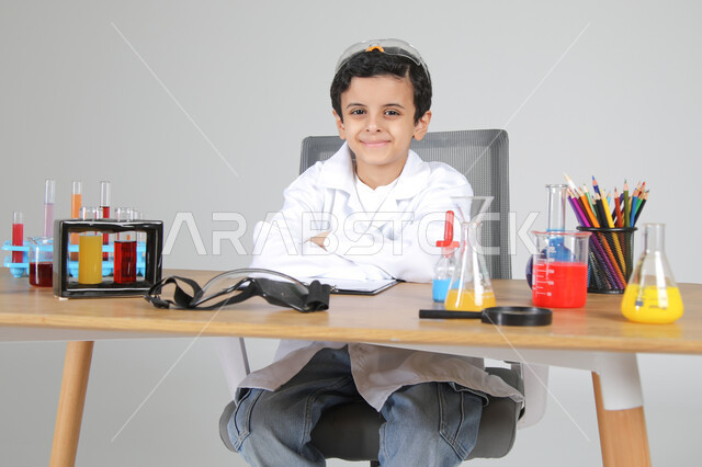 تجسيد مهن الأطفال المستقبلية منذ الصغر، شغف الوصول إلى حلم دراسة الصيدلة مستقبلا، التجارب الكيميائية للعلماء الصغار، بورتريه مقرب لصبي عربي خليجي سعودي يرتدي المعطف الطبي يجلس خلف المكتب وأمامه زجاجات التحاليل وينظر إلى الكاميرا بإيماءات السرور، خلفية رمادية