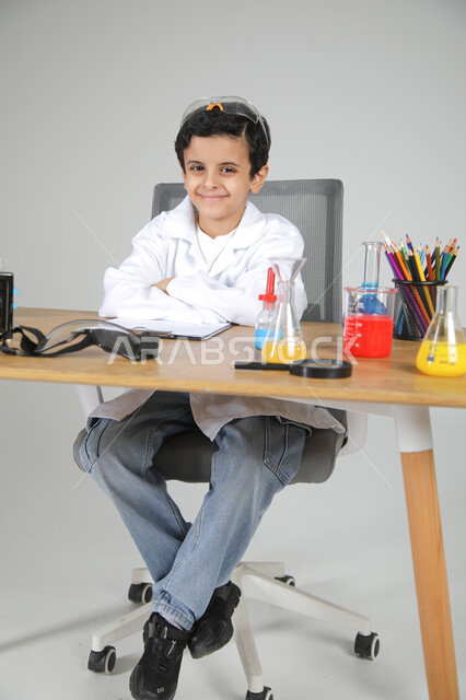 شغف الوصول إلى حلم دراسة الصيدلة مستقبلا، التجارب الكيميائية للعلماء الصغار، تجسيد مهن الأطفال المستقبلية منذ الصغر، بورتريه لصبي عربي خليجي سعودي يرتدي المعطف الطبي يجلس وأمامه زجاجات التحاليل وينظر للكاميرا بإيماءات السرور، الجسم بالطول الكامل، خلفية رمادية