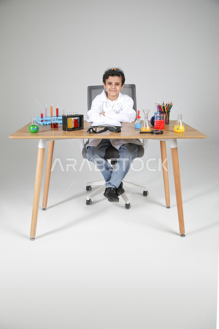 شغف الوصول إلى حلم دراسة الصيدلة مستقبلا، التجارب الكيميائية للعلماء الصغار، تجسيد مهن الأطفال المستقبلية منذ الصغر، بورتريه لصبي عربي خليجي سعودي يرتدي المعطف الطبي يجلس وأمامه زجاجات التحاليل وينظر للكاميرا بإيماءات السرور، الجسم بالطول الكامل، خلفية رمادية