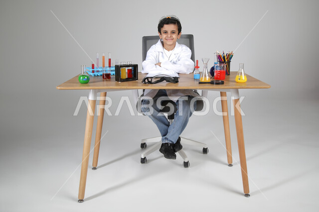 شغف الوصول إلى حلم دراسة الصيدلة مستقبلا، التجارب الكيميائية للعلماء الصغار، تجسيد مهن الأطفال المستقبلية منذ الصغر، بورتريه لصبي عربي خليجي سعودي يرتدي المعطف الطبي يجلس وأمامه زجاجات التحاليل وينظر للكاميرا بإيماءات السرور، الجسم بالطول الكامل، خلفية رمادية