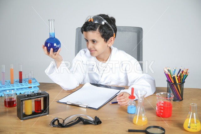التجارب والأبحاث الكيميائية للعلماء الصغار، شغف الوصول إلى حلم دراسة الصيدلة مستقبلا، تجسيد مهن الأطفال المستقبلية منذ الصغر، بورتريه مقرب لصبي عربي خليجي سعودي يرتدي المعطف الطبي يجلس خلف المكتب ويمسك بزجاجات التحاليل ويقوم بتسجيل الملاحظات، خلفية رمادية