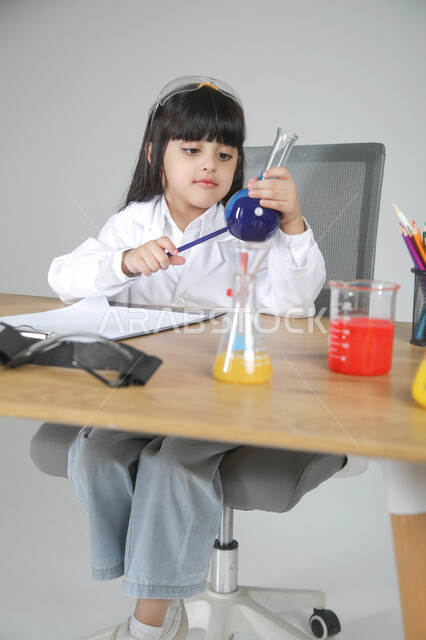 تفحص زجاجة التحليل وقياس التراكيز بدقة، التجارب الكيميائية للعلماء الصغار، شغف الوصول إلى حلم دراسة الصيدلة مستقبلا، تجسيد مهن الأطفال المستقبلية منذ الصغر، بورتريه لبنت عربية خليجية سعودية ترتدي المعطف الطبي تجلس خلف المكتب وتسجل الملاحظات، خلفية رمادية