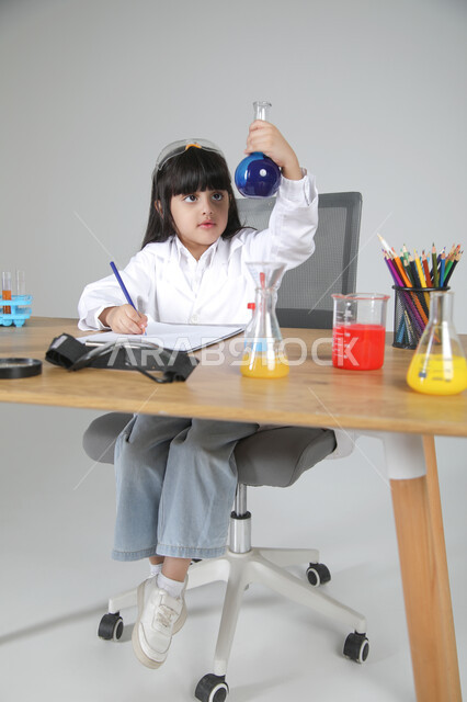 النظر إلى زجاجة التحليل وتفحصها، التجارب الكيميائية للعلماء الصغار، شغف الوصول إلى حلم دراسة الصيدلة مستقبلا، تجسيد مهن الأطفال المستقبلية منذ الصغر، بورتريه لبنت عربية خليجية سعودية ترتدي المعطف الطبي تجلس خلف المكتب، الجسم بالطول الكامل، خلفية رمادية