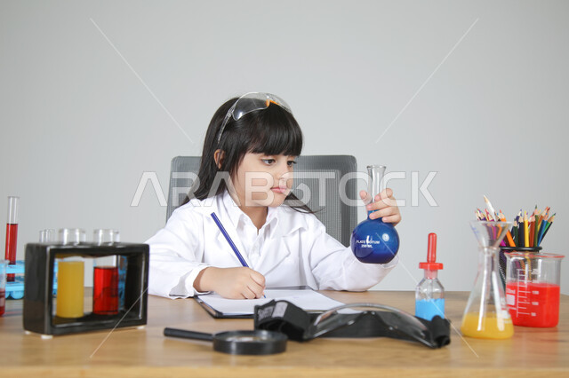 تسجيل الملاحظات بإيماءات التركيز، التجارب الكيميائية للعلماء الصغار، شغف الوصول إلى حلم دراسة الصيدلة مستقبلا، تجسيد مهن الأطفال المستقبلية منذ الصغر، بورتريه مقرب لبنت عربية خليجية سعودية ترتدي المعطف الطبي تجلس وتمسك بزجاجات التحاليل الملونة، خلفية رمادية