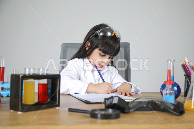 تسجيل الملاحظات حول التحاليل والتراكيز بإيماءات التركيز، التجارب الكيميائية للعلماء الصغار، شغف الوصول إلى حلم دراسة الصيدلة مستقبلا، تجسيد مهن الأطفال المستقبلية منذ الصغر، بورتريه مقرب لبنت عربية خليجية سعودية ترتدي المعطف الطبي تجلس خلف المكتب، خلفية رمادية