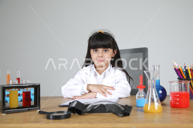  النظر إلى الكاميرا بإيماءات السرور، التجارب الكيميائية للعلماء الصغار، شغف الوصول إلى حلم دراسة الصيدلة مستقبلا، تجسيد مهن الأطفال المستقبلية منذ الصغر، بورتريه مقرب لبنت عربية خليجية سعودية ترتدي المعطف الطبي تجلس خلف المكتب بثقة بالنفس، خلفية رمادية