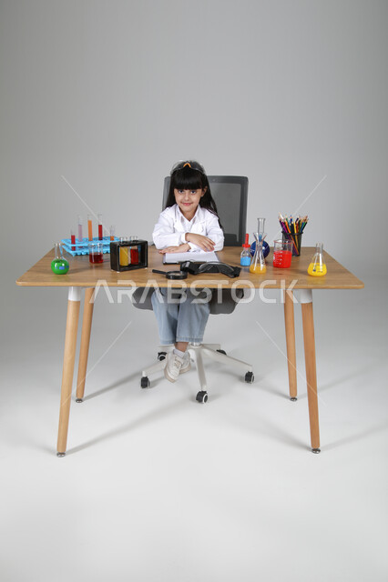 النظر إلى الكاميرا بإيماءات السرور، التجارب الكيميائية للعلماء الصغار، شغف الوصول إلى حلم دراسة الصيدلة مستقبلا، تجسيد مهن الأطفال المستقبلية منذ الصغر، بورتريه لبنت عربية خليجية سعودية ترتدي المعطف الطبي تجلس خلف المكتب، الجسم بالطول الكامل، خلفية رمادية