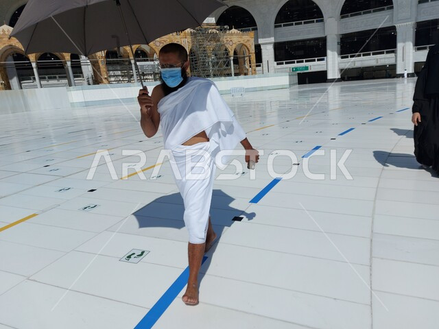 أماكن العبادة والتقرب الى الله, أداء مناسك الحج والعمرة والوقاية من فايروس كورونا، صورة مقربة لرجل مسلم خليجي عربي سعودي يرتدي الكمامة الواقية و ملابس الاحرام يحمل بيده مظلة رمادية في مكة المكرمة، صورة الجسم بالطول الكامل