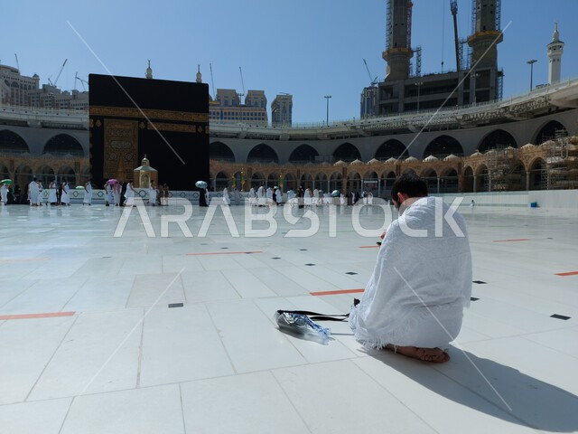 أداء مناسك الحج والعمرة، العبادة والتقرب إلى الله، طواف الحجاج والمعتمرين حول الكعبة المشرفة، صورة مقربة من الخلف لرجل مسلم عربي خليجي سعودي يرتدي لباس الإحرام  بجلس في ساحة الحرم المكي الشريف في مكة المكرمة، أماكن اسلامية دينية مقدسة في السعودية
