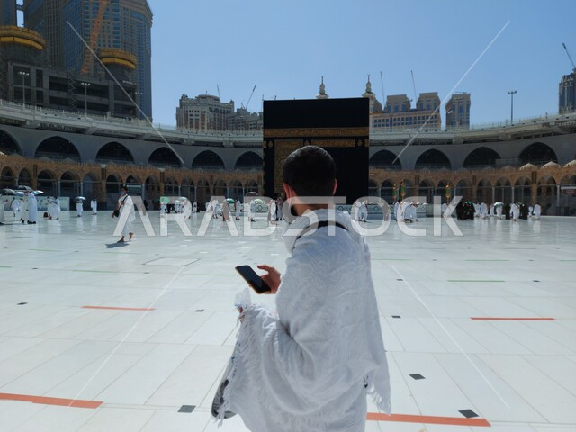 مفهوم العبادة والطاعة والتقرب الى الله، المعتمرون يطوفون حول الكعبه، مناسك العمرة في مكة المكرمة أيام انتشار فايروس كورونا في السعودية، صورة مقربة من الخلف لرجل مسلم يرتدي لباس الاحرام يمسك بيده الهاتف المحمول، التواصل مع الاهل والاصدقاء، أماكن دينية مقدسة