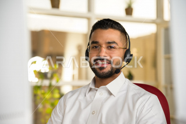 موظف عربي خليجي سعودي يعمل في خدمة العملاء ويرتدي سماعة الاتصال، استعلامات واستفسارات، الاجابه على اسئلة العملاء، اجراء اتصال فيديو من خلال الحاسوب، نظارة طبية، متابعة وتطوير العمل