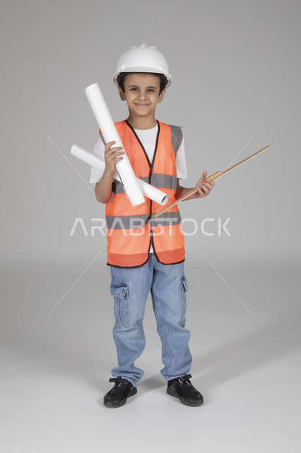 الإمساك بمخططات الإعمار وأدوات القياس الهندسية، تجريب الأطفال للمهن المستقبلية منذ الصغر، تجسيد تجربة العمل في المجال الهندسي، بورتريه لولد عربي خليجي سعودي يرتدي سترة وخوذة الحماية يقف وينظر إلى الكاميرا بإيماءات السعادة، الجسم بالطول الكامل، خلفية رماية