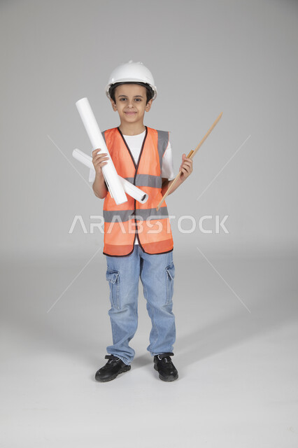 Holding construction plans and engineering measuring tools, children experimenting with future professions from an early age, embodying the experience of working in the engineering field, portrait of a Saudi Arabian Gulf boy wearing a protective jacket and helmet standing and looking at the camera with gestures of happiness, full-length body, shooting background