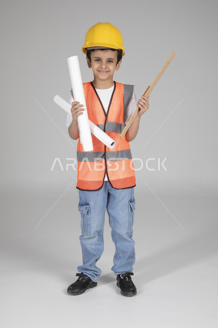 Holding construction plans and engineering measuring tools, children experimenting with future professions from an early age, embodying the experience of working in the engineering field, portrait of a Saudi Arabian Gulf boy wearing a protective jacket and helmet standing and looking at the camera with gestures of happiness, full-length body, shooting background