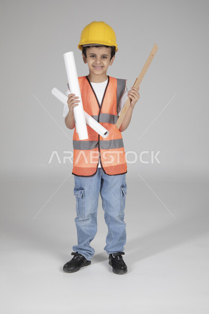 الإمساك بمخططات الإعمار وأدوات القياس الهندسية، تجريب الأطفال للمهن المستقبلية منذ الصغر، تجسيد تجربة العمل في المجال الهندسي، بورتريه لولد عربي خليجي سعودي يرتدي سترة وخوذة الحماية يقف وينظر إلى الكاميرا بإيماءات السعادة، الجسم بالطول الكامل، خلفية رماية