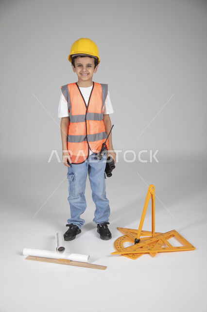 الوقوف والنظر إلى الكاميرا بإيماءات السعادة، تجريب الأطفال للمهن المستقبلية منذ الصغر، تجسيد تجربة العمل في المجال الهندسي، بورتريه لولد عربي خليجي سعودي يرتدي سترة وخوذة الحماية، مخطط الإعمار والأدوات الهندسية والمنظار المقرب، الجسم بالطول الكامل، خلفية رماية