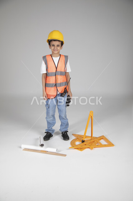 الوقوف والنظر إلى الكاميرا بإيماءات السعادة، تجريب الأطفال للمهن المستقبلية منذ الصغر، تجسيد تجربة العمل في المجال الهندسي، بورتريه لولد عربي خليجي سعودي يرتدي سترة وخوذة الحماية، مخطط الإعمار والأدوات الهندسية والمنظار المقرب، الجسم بالطول الكامل، خلفية رماية