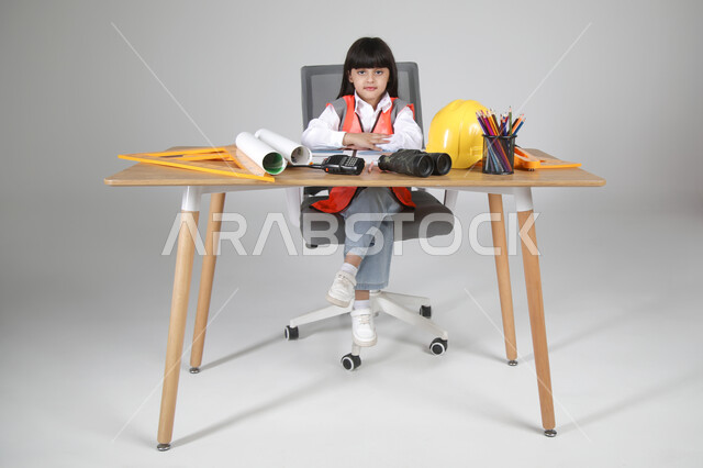 النظر للكاميرا بإيماءات البشاشة، تجريب الأطفال للمهن المستقبلية منذ الصغر، تجسيد تجربة العمل كمهندسة، بورتريه لبنت عربية خليجية سعودية ترتدي سترة الحماية تجلس خلف المكتب وأمامها مخطط الإعمار والأدوات الهندسية والمنظار والخوذة، الجسم بالطول الكامل، خلفية رماية