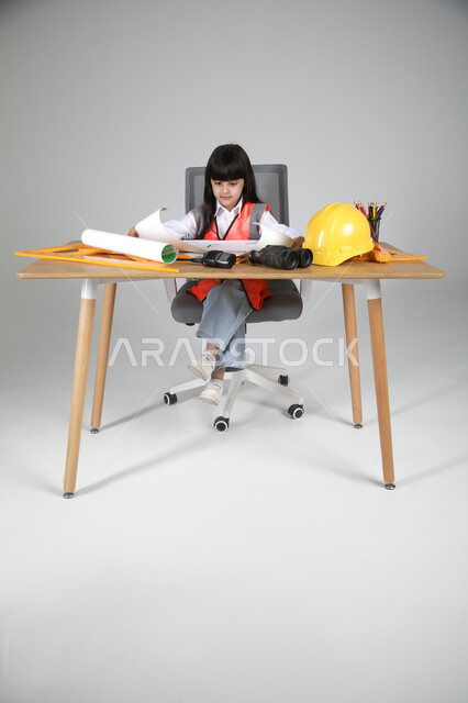 تجسيد تجربة العمل كمهندسة معمارية، تجريب الأطفال للمهن المستقبلية منذ الصغر، بورتريه لبنت عربية خليجية سعودية ترتدي سترة الحماية تجلس خلف المكتب وتتابع مخطط الإعمار بإيماءات التركيز، الأدوات الهندسية والمنظار والخوذة الصفراء، الجسم بالطول الكامل، خلفية رماية