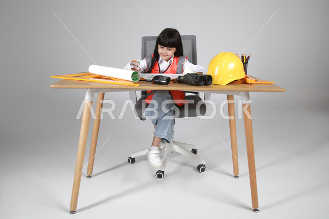 تجسيد تجربة العمل كمهندسة معمارية، تجريب الأطفال للمهن المستقبلية منذ الصغر، بورتريه لبنت عربية خليجية سعودية ترتدي سترة الحماية تجلس خلف المكتب وتتابع مخطط الإعمار بإيماءات التركيز، الأدوات الهندسية والمنظار والخوذة الصفراء، الجسم بالطول الكامل، خلفية رماية