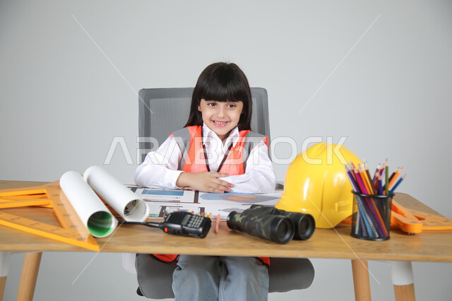 فرحة الأطفال في تجريب المهن المستقبلية منذ الصغر، تجسيد تجربة العمل في المجال الهندسي، بورتريه مقرب لبنت عربية خليجية سعودية ترتدي سترة الحماية تجلس خلف المكتب وأمامها مخططات الإعمار تنظر للكاميرا بسرور، استخدام الأدوات الهندسية والمنظار والخوذة، خلفية رماية