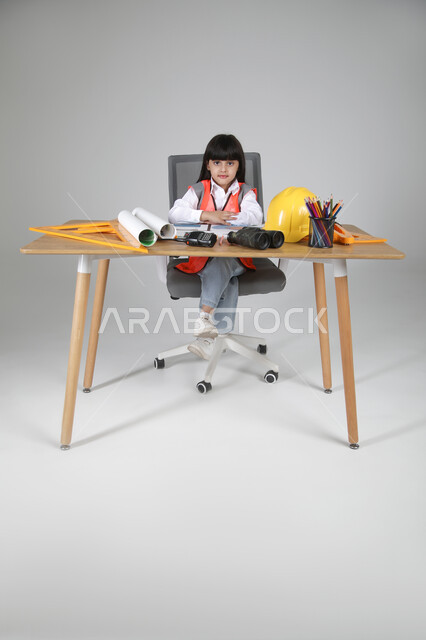 النظر للكاميرا بإيماءات البشاشة، تجريب الأطفال للمهن المستقبلية منذ الصغر، تجسيد تجربة العمل كمهندسة، بورتريه لبنت عربية خليجية سعودية ترتدي سترة الحماية تجلس خلف المكتب وأمامها مخطط الإعمار والأدوات الهندسية والمنظار والخوذة، الجسم بالطول الكامل، خلفية رماية