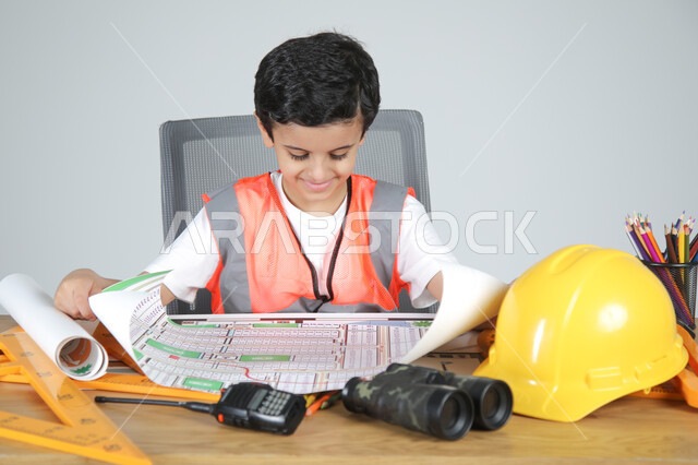 استمتاع الأطفال في تجريب المهن المستقبلية منذ الصغر، تجسيد تجربة العمل في المجال الهندسي، بورتريه مقرب لولد عربي خليجي سعودي يرتدي سترة الحماية يجلس خلف المكتب ويمسك مخططات الإعمار ويقوم بمراجعتها، استخدام الأدوات الهندسية والمنظار والخوذة، خلفية رماية