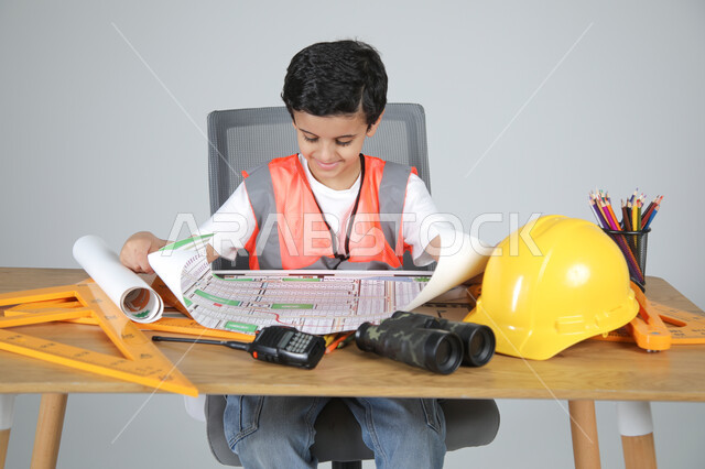 استمتاع الأطفال في تجريب المهن المستقبلية منذ الصغر، تجسيد تجربة العمل في المجال الهندسي، بورتريه مقرب لولد عربي خليجي سعودي يرتدي سترة الحماية يجلس خلف المكتب ويمسك مخططات الإعمار ويقوم بمراجعتها، استخدام الأدوات الهندسية والمنظار والخوذة، خلفية رماية