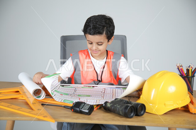 استمتاع الأطفال في تجريب المهن المستقبلية منذ الصغر، تجسيد تجربة العمل في المجال الهندسي، بورتريه مقرب لولد عربي خليجي سعودي يرتدي سترة الحماية يجلس خلف المكتب ويمسك مخططات الإعمار ويقوم بمراجعتها، استخدام الأدوات الهندسية والمنظار والخوذة، خلفية رماية