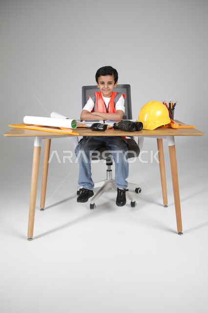 Gestures of happiness in trying out future professions since childhood, embodying the experience of children working in the engineering field, portrait of a Saudi Arabian Gulf boy wearing a protective vest sitting behind a desk with construction plans in front of him, using engineering tools, binoculars and helmet, full-length body, shooting background