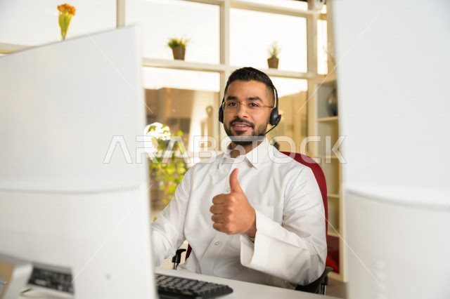 A Saudi Arabian Gulf employee working in customer service, raising his thumb up, wearing a calling headset, answering customer questions, making a video call through a computer, eyeglasses, facial and hand gestures indicating approval and admiration.