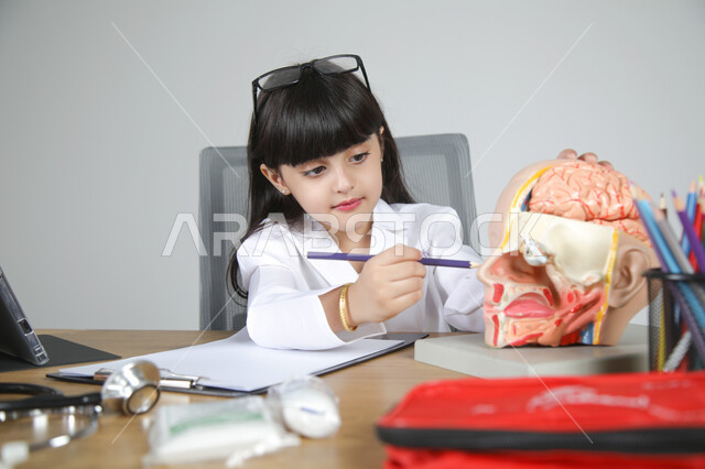 استخدام المجسمات التشريحية، اختيار مهنة المستقبل منذ الصغر، استمتاع الأطفال بتجربة العمل بالمجال الصحي، بورتريه مقرب لفتاة عربية خليجية سعودية ترتدي المعطف الطبي وتضع سماعة الفحص حول عنقها تجلس خلف المكتب تمسك القلم وتشير لعضو معين في جسم الإنسان، خلفية رمادية