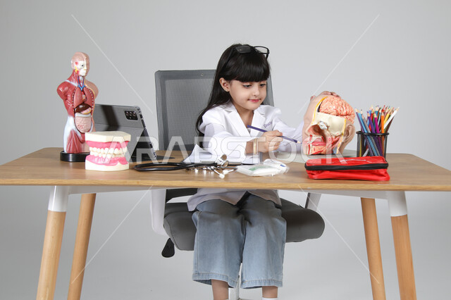 استخدام المجسمات التشريحية، اختيار مهنة المستقبل منذ الصغر، استمتاع الأطفال بتجربة العمل بالمجال الصحي، بورتريه مقرب لفتاة عربية خليجية سعودية ترتدي المعطف الطبي وتضع سماعة الفحص حول عنقها تجلس خلف المكتب تمسك القلم وتشير لعضو معين في جسم الإنسان، خلفية رمادية