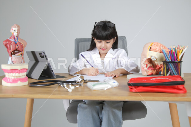 تحديد و اختيار مهنة المستقبل منذ الصغر، استمتاع الأطفال بتجربة العمل بمجال الرعاية الصحية، بورتريه لفتاة عربية خليجية سعودية ترتدي المعطف الطبي وتضع سماعة الفحص حول عنقها تجلس خلف المكتب تقوم بتسجيل الملاحظات بإيماءات التركيز، الجسم بالطول الكامل، خلفية رمادية