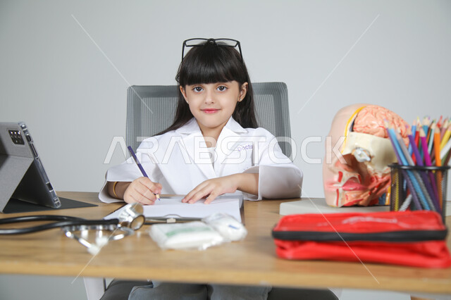 النظر إلى الكاميرا بإيماءات البشاشة، اختيار مهنة المستقبل منذ الصغر، استمتاع الأطفال بتجربة العمل في المجال الصحي، بورتريه مقرب لفتاة عربية خليجية سعودية ترتدي المعطف الطبي وتضع سماعة الفحص حول عنقها تجلس خلف المكتب وأمامها المجسمات التشريحية، خلفية رمادية