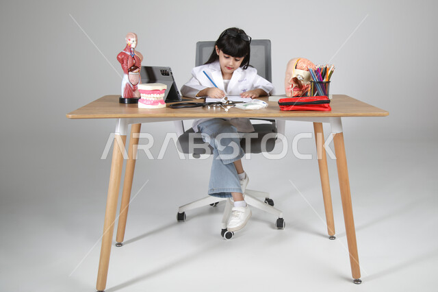 تحديد و اختيار مهنة المستقبل منذ الصغر، استمتاع الأطفال بتجربة العمل بمجال الرعاية الصحية، بورتريه لفتاة عربية خليجية سعودية ترتدي المعطف الطبي وتضع سماعة الفحص حول عنقها تجلس خلف المكتب تقوم بتسجيل الملاحظات بإيماءات التركيز، الجسم بالطول الكامل، خلفية رمادية