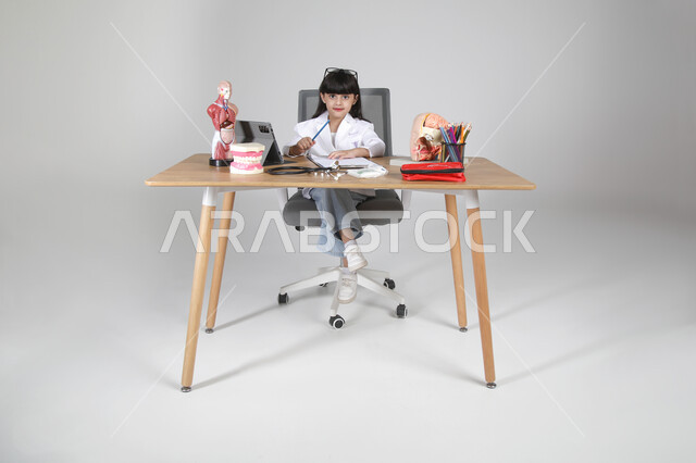 تحديد و اختيار مهنة المستقبل منذ الصغر، استمتاع الأطفال بتجربة العمل بمجال الرعاية الصحية، بورتريه لفتاة عربية خليجية سعودية ترتدي المعطف الطبي وتضع سماعة الفحص حول عنقها تجلس خلف المكتب تنظر إلى الكاميرا بإيماءات السعادة، الجسم بالطول الكامل، خلفية رمادية