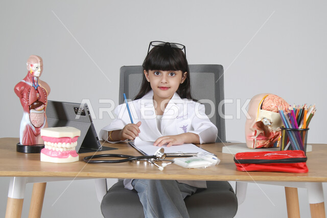 النظر إلى الكاميرا بإيماءات البشاشة، اختيار مهنة المستقبل منذ الصغر، استمتاع الأطفال بتجربة العمل في المجال الصحي، بورتريه مقرب لفتاة عربية خليجية سعودية ترتدي المعطف الطبي وتضع سماعة الفحص حول عنقها تجلس خلف المكتب وأمامها المجسمات التشريحية، خلفية رمادية