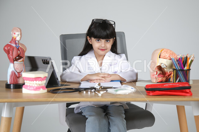 النظر إلى الكاميرا بإيماءات البشاشة، اختيار مهنة المستقبل منذ الصغر، استمتاع الأطفال بتجربة العمل في المجال الصحي، بورتريه مقرب لفتاة عربية خليجية سعودية ترتدي المعطف الطبي وتضع سماعة الفحص حول عنقها تجلس خلف المكتب وأمامها المجسمات التشريحية، خلفية رمادية