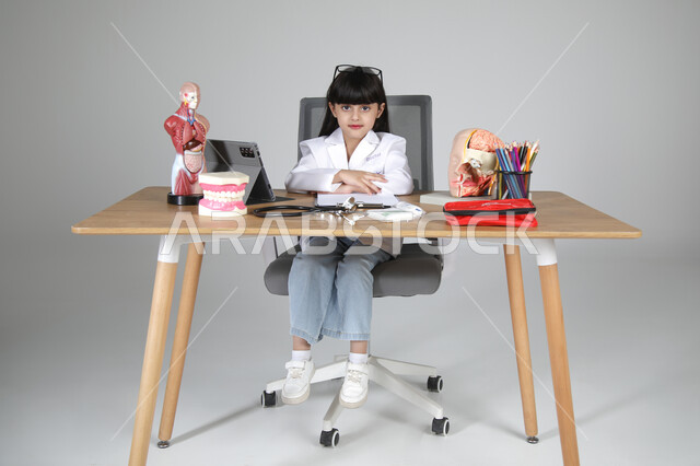 Determining and choosing a future career from an early age, children enjoying the experience of working in the healthcare field, portrait of a Saudi Arabian Gulf girl wearing a medical coat and a stethoscope around her neck sitting behind a desk looking at the camera with gestures of happiness, full body, gray background