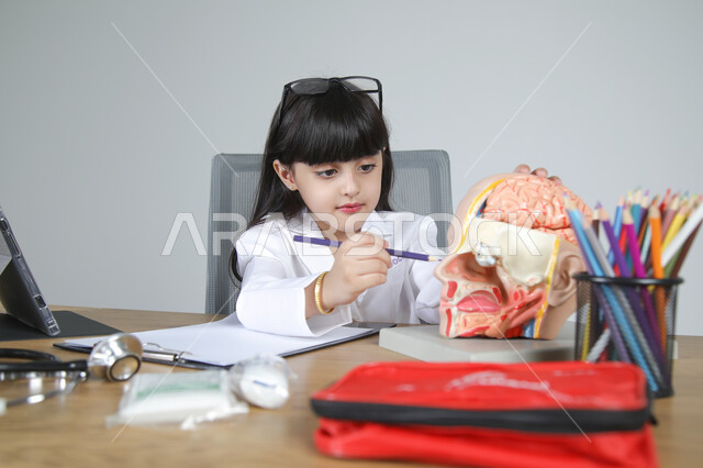 استخدام المجسمات التشريحية، اختيار مهنة المستقبل منذ الصغر، استمتاع الأطفال بتجربة العمل بالمجال الصحي، بورتريه مقرب لفتاة عربية خليجية سعودية ترتدي المعطف الطبي وتضع سماعة الفحص حول عنقها تجلس خلف المكتب تمسك القلم وتشير لعضو معين في جسم الإنسان، خلفية رمادية