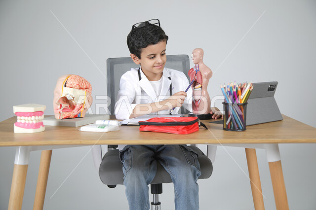 استخدام المجسمات التشريحية، اختيار مهنة المستقبل منذ الصغر، استمتاع الأطفال بتجربة العمل في المجال الصحي، بورتريه مقرب لولد عربي خليجي سعودي يرتدي المعطف الطبي ويضع سماعة الفحص حول عنقه يجلس خلف المكتب يمسك القلم ويشير إلى عضو معين في جسم الإنسان، خلفية رمادية