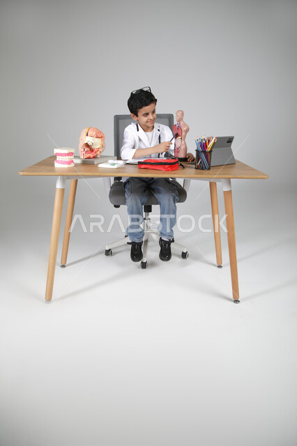 الإشارة إلى أعضاء الجسم باستخدام المجسمات التشريحية، اختيار مهنة المستقبل منذ الصغر، استمتاع الأطفال بتجربة العمل بالمجال الصحي، بورتريه لولد عربي خليجي سعودي يرتدي المعطف الطبي ويضع سماعة الفحص حول عنقه ويجلس خلف طاولة المكتب، الجسم بالطول الكامل، خلفية رمادية