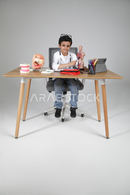 الإشارة إلى أعضاء الجسم باستخدام المجسمات التشريحية، اختيار مهنة المستقبل منذ الصغر، استمتاع الأطفال بتجربة العمل بالمجال الصحي، بورتريه لولد عربي خليجي سعودي يرتدي المعطف الطبي ويضع سماعة الفحص حول عنقه ويجلس خلف طاولة المكتب، الجسم بالطول الكامل، خلفية رمادية