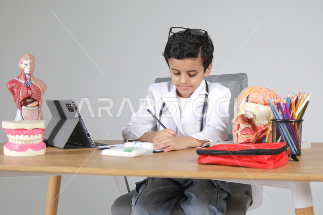 اختيار مهنة المستقبل منذ الصغر، استمتاع الأطفال بتجربة العمل في المجال الصحي، بورتريه مقرب لولد عربي خليجي سعودي يرتدي المعطف الطبي ويضع سماعة الفحص حول عنقه يجلس خلف المكتب ويسجل الملاحظات بإيماءات التركيز، المجسمات التشريحية لأعضاء جسم الإنسان، خلفية رمادية