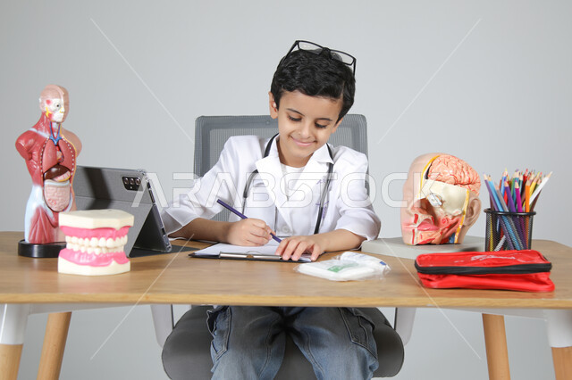 اختيار مهنة المستقبل منذ الصغر، استمتاع الأطفال بتجربة العمل في المجال الصحي، بورتريه مقرب لولد عربي خليجي سعودي يرتدي المعطف الطبي ويضع سماعة الفحص حول عنقه يجلس خلف المكتب ويسجل الملاحظات بإيماءات التركيز، المجسمات التشريحية لأعضاء جسم الإنسان، خلفية رمادية