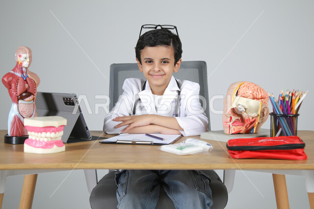 اختيار مهنة المستقبل منذ الصغر، استمتاع الأطفال بتجربة العمل في المجال الصحي، بورتريه مقرب لولد عربي خليجي سعودي يرتدي المعطف الطبي ويضع سماعة الفحص حول عنقه يجلس خلف المكتب وينظر للكاميرا بإيماءات السعادة، المجسمات التشريحية لأعضاء جسم الإنسان، خلفية رمادية