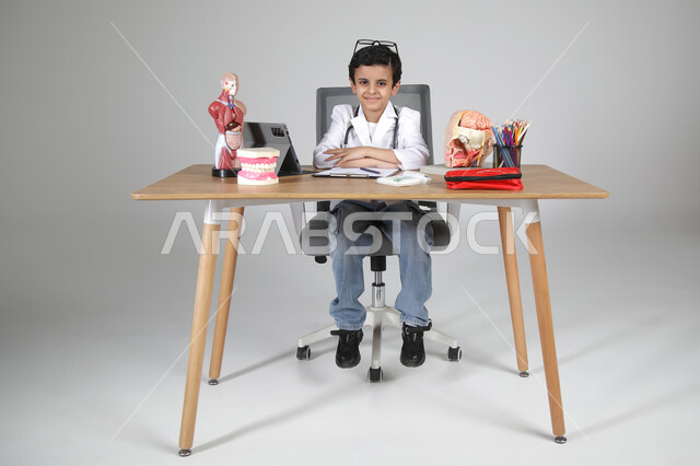 النظر إلى الكاميرا بإيماءات السرور، اختيار مهنة المستقبل منذ الصغر، استمتاع الأطفال بتجربة العمل بالمجال الصحي، بورتريه لولد عربي خليجي سعودي يرتدي المعطف الطبي ويضع سماعة الفحص حول عنقه ويجلس خلف المكتب وأمامه مجسمات تشريحية، الجسم بالطول الكامل، خلفية رمادية