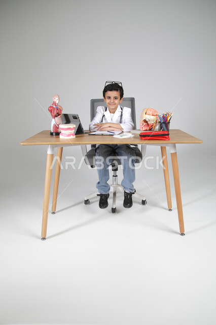 النظر إلى الكاميرا بإيماءات السرور، اختيار مهنة المستقبل منذ الصغر، استمتاع الأطفال بتجربة العمل بالمجال الصحي، بورتريه لولد عربي خليجي سعودي يرتدي المعطف الطبي ويضع سماعة الفحص حول عنقه ويجلس خلف المكتب وأمامه مجسمات تشريحية، الجسم بالطول الكامل، خلفية رمادية